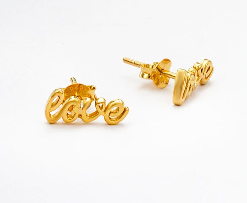 May include: A pair of gold-coloured stud earrings. Each earring is shaped to spell out the word "love" in a cursive font. The earrings are set against a white background.