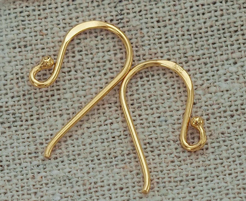Op de afbeelding: Een paar goudkleurige metalen oorhaken met een glad, gebogen ontwerp. Elke haak is voorzien van een klein lusje aan de onderkant voor het bevestigen van sieradenonderdelen en een afgerond uiteinde. Het metaal heeft een gepolijste afwerking.