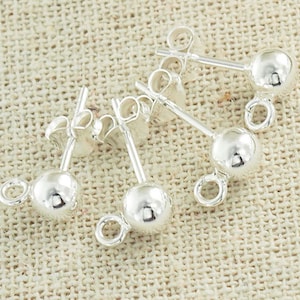 Può includere: Primo piano di tre paia di orecchini a bottone in argento. Ogni orecchino presenta una piccola sfera lucida alla fine di un perno, con un anello attaccato. Gli orecchini sono esposti su una superficie beige strutturata.