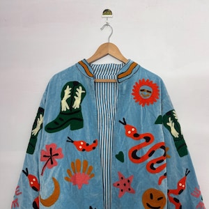 May include: Light blue jacket with embroidered designs. Features a green cowboy boot, a smiling sun, and red snakes. The jacket has a striped lining and is hanging on a wooden hanger. The jacket is decorated with various colorful embroidered shapes.