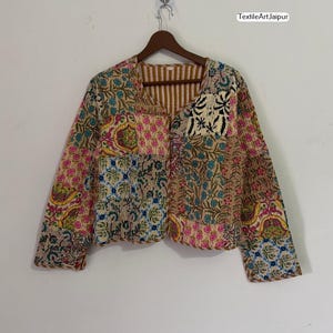 Könnte beinhalten: Patchwork-Jacke mit verschiedenen Blumen- und Paisley-Mustern in Rosa, Blau, Gelb und Braun. Die Jacke hat ein gestreiftes Futter und hängt an einem Holzbügel.