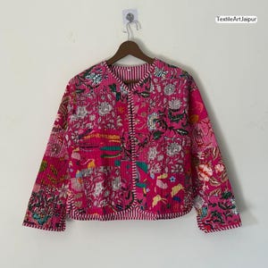 May include: A vibrant pink jacket with a floral pattern and white striped trim. The jacket is displayed on a wooden hanger against a white wall. The text "TextileArtJaipur" is visible.