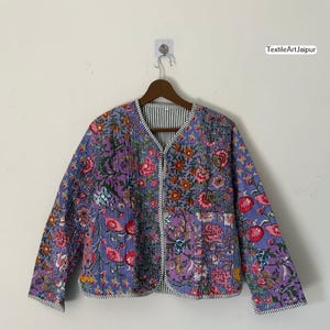 May include: A quilted jacket with a floral design in purple, pink, and orange hues. The jacket features a V-neck and a striped lining. It is displayed on a wooden hanger. The jacket's measurements are in centimeters.