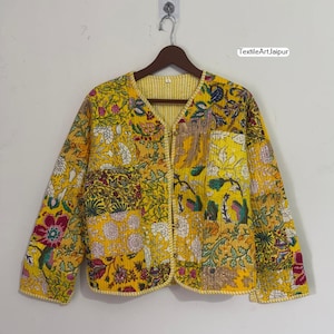 May include: A yellow quilted jacket with a floral patchwork design. The jacket has a V-neck and long sleeves. The jacket is hanging on a wooden hanger against a white wall. The text "TextileArtJaipur" is visible.