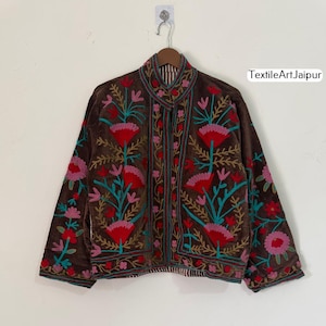 Puede incluir: Una chaqueta de terciopelo marrón con bordados florales coloridos. La chaqueta presenta diseños florales rojos, rosas y turquesas. El texto "TextileArt Jaipur" es visible en la parte superior derecha.