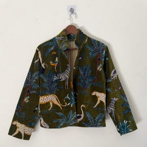 May include: An olive green jacket with a jungle pattern featuring leopards, zebras, birds, and tropical plants. The jacket has a mandarin collar and long sleeves. The jacket is hung on a wooden hanger.