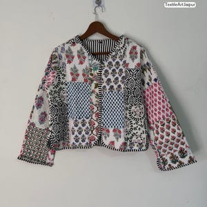 Peut inclure: Veste matelassée avec un motif patchwork. La veste présente divers motifs, notamment floraux, géométriques et à pois, dans des tons de rouge, bleu, noir et blanc. Le texte "TextileArtJaipur" est visible. La veste est suspendue à un cintre en bois.