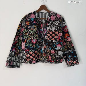 Puede incluir: Chaqueta acolchada negra con un estampado floral en rosa, rojo, verde y blanco. La chaqueta tiene forro y ribete a rayas. El texto "TextileArtJaipur" es visible en la parte superior derecha.