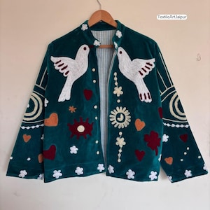 May include: A teal velvet jacket with white dove and floral embroidery. The jacket features a striped lining and is decorated with embroidered hearts, flowers, and sun motifs. The text "TextileArtJaipur" is visible on the upper right.