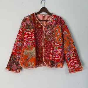 Puede incluir: Chaqueta patchwork en tonos rojos, naranjas y marrones, con estampados florales y geométricos. La chaqueta tiene cuello en V, mangas largas y forro a rayas. La chaqueta está colgada en una percha de madera.