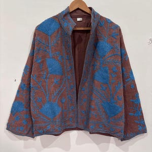 Può includere: Giacca marrone con ricami floreali blu. La giacca aperta è appesa a una gruccia di legno. Il ricamo presenta fiori e foglie stilizzate, creando un motivo decorativo. L'interno della giacca è di colore marrone pieno.