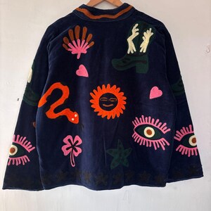 May include: Navy blue jacket with colorful embroidered designs. The jacket features a variety of embroidered images, including a snake, sun, eyes, hearts, and a cowboy boot. The jacket is hanging on a wooden hanger.
