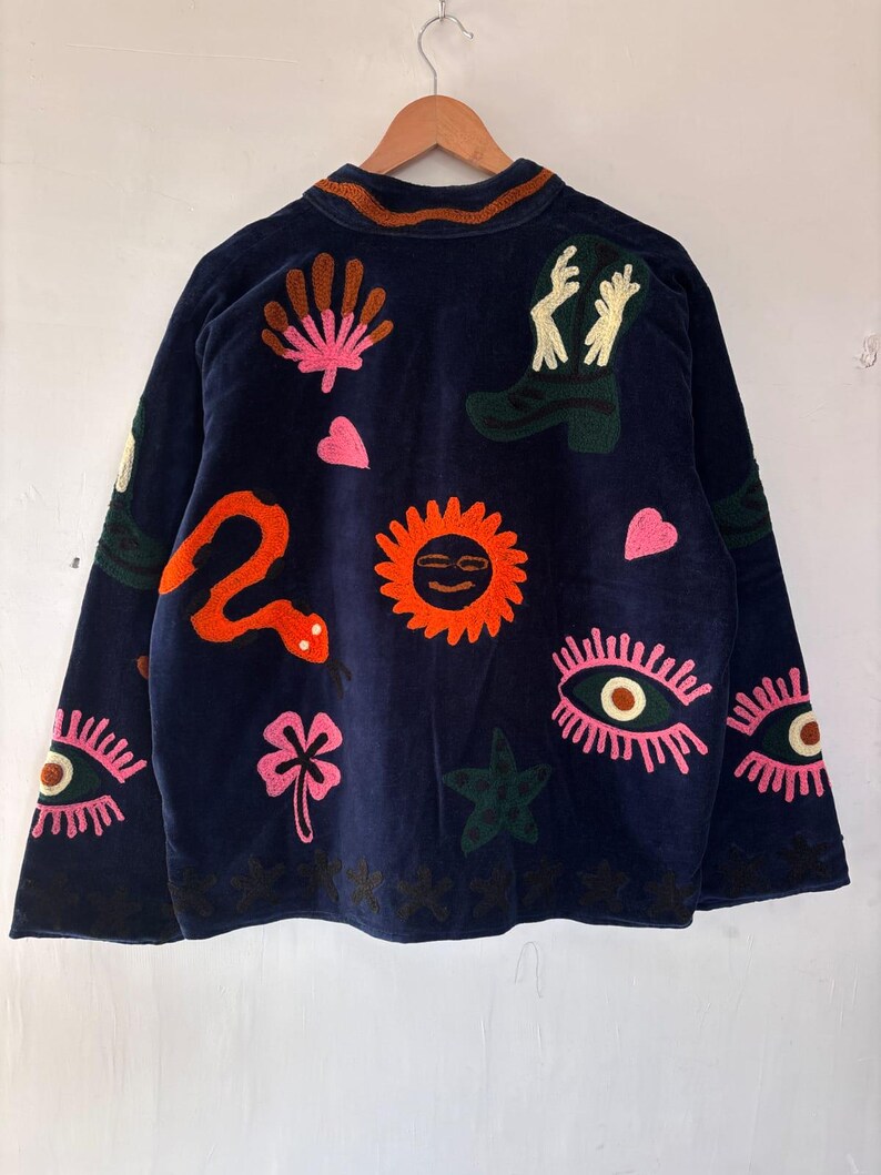 May include: Navy blue jacket with colorful embroidered designs. The jacket features a variety of embroidered images, including a snake, sun, eyes, hearts, and a cowboy boot. The jacket is hanging on a wooden hanger.