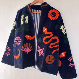 May include: A navy blue jacket with colorful embroidered designs, including a sun, snake, and various floral and abstract shapes. The jacket has a striped lining and is hanging on a wooden hanger.