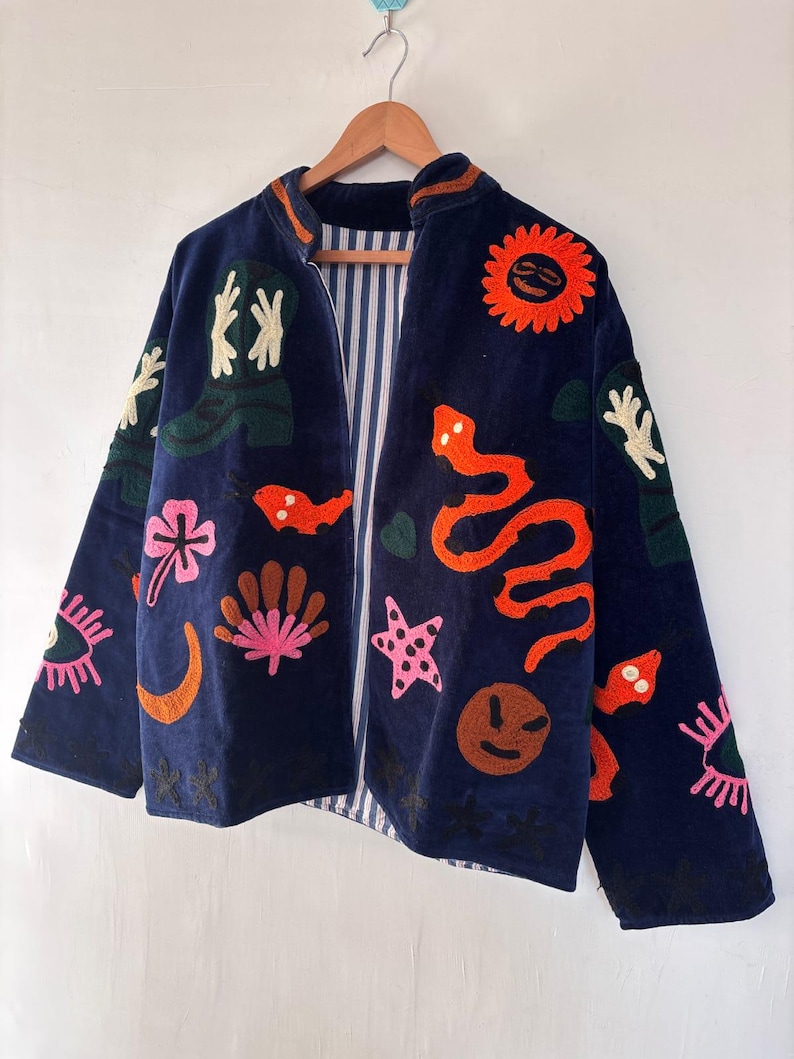 May include: A navy blue jacket with colorful embroidered designs, including a sun, snake, and various floral and abstract shapes. The jacket has a striped lining and is hanging on a wooden hanger.