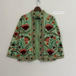 May include: A pale green jacket featuring intricate floral embroidery in red, pink, and orange. The jacket, with a mandarin collar, is displayed on a wooden hanger. The text "TextileArt Jaipur" is visible on the right side.