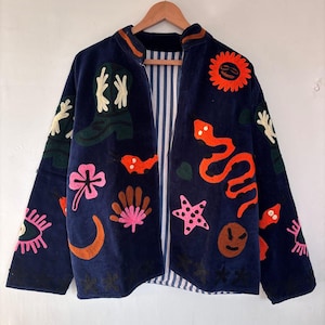 May include: Navy blue jacket with colorful embroidered designs. Features a variety of motifs including a sun, snake, boots, and flowers. The jacket has a striped lining and is hanging on a wooden hanger.