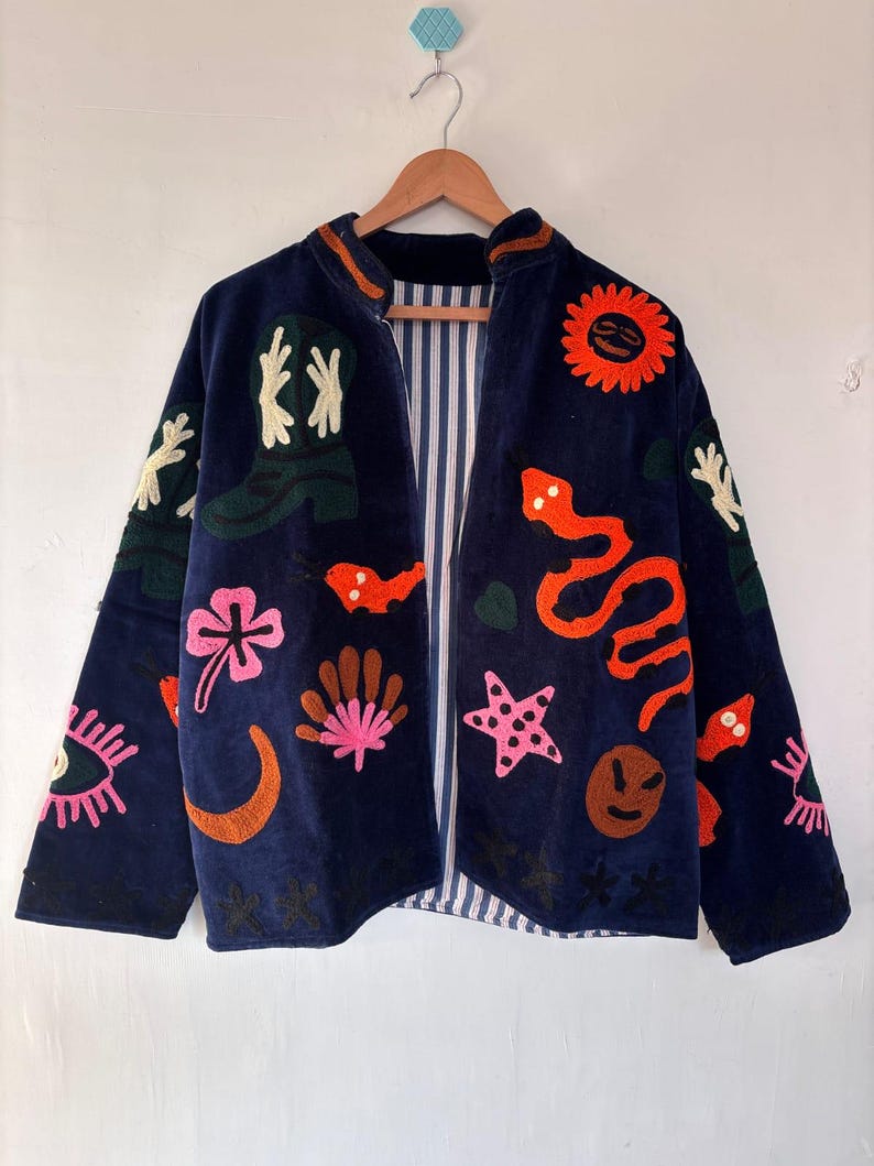 May include: Navy blue jacket with colorful embroidered designs. Features a variety of motifs including a sun, snake, boots, and flowers. The jacket has a striped lining and is hanging on a wooden hanger.