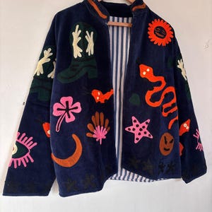 May include: Navy blue jacket with colorful embroidered designs, including a snake, stars, and a sun. The jacket has a striped lining and is hanging on a wooden hanger.