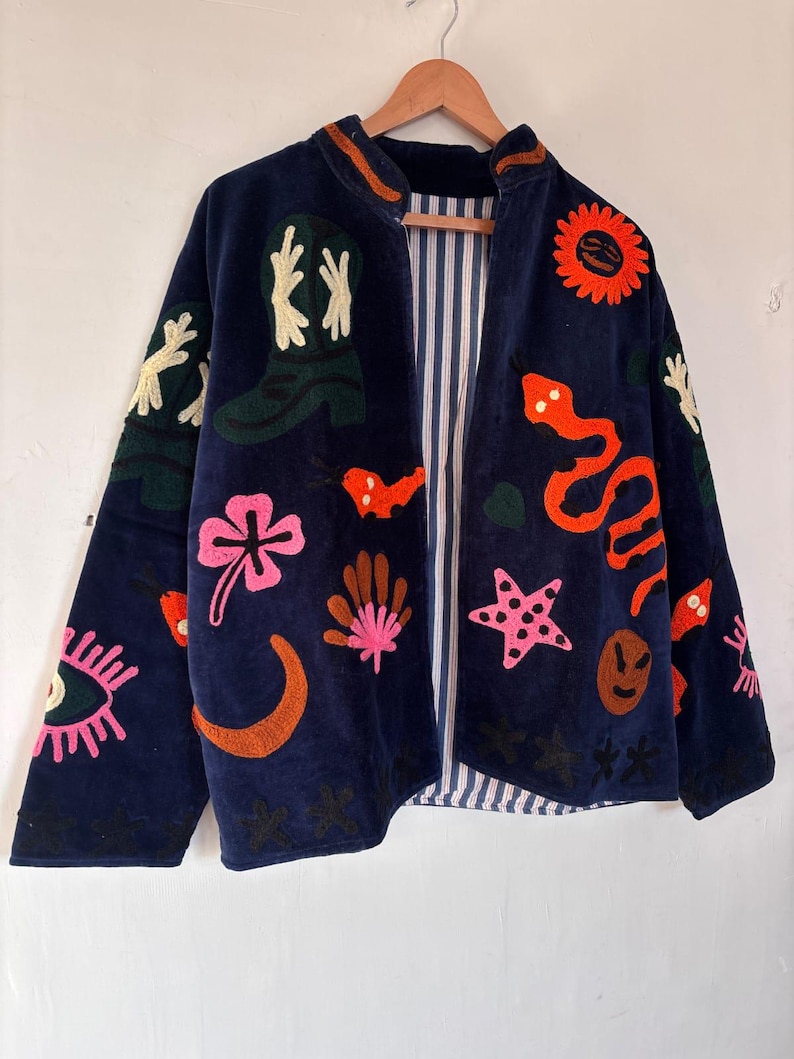 May include: Navy blue jacket with colorful embroidered designs, including a snake, stars, and a sun. The jacket has a striped lining and is hanging on a wooden hanger.