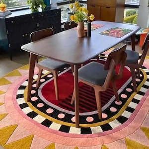 May include: A dining table and chairs set on a patterned rug. The table is dark wood, with four chairs with grey seats. The rug is oval, with a red centre and a border of pink, yellow, and black geometric shapes.