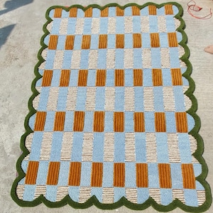 May include: A rectangular rug with a checkered pattern in shades of blue, tan, and brown. The rug has a scalloped green border. The design features alternating vertical stripes of blue, tan, and brown, creating a visually appealing geometric pattern.