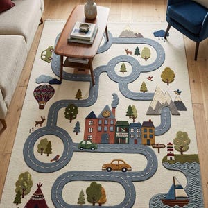 May include: A cream-colored children's rug with a road design featuring cars, buildings, trees, mountains, and a hot air balloon. The rug has a whimsical design with a lighthouse, a teepee, and a sailboat. The word "LIBRARY" is visible on one of the buildings.
