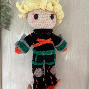 K&ouml;nnte beinhalten: Handgefertigte H&auml;kelpuppe mit einem Charakter mit stacheligen blonden Haaren, einem schwarzen Outfit mit orangefarbenen und gr&uuml;nen Akzenten und grauen Knieschonern. Die Puppe hat einen hellen Hautton und rote Augen. Die Puppe ist etwa 25 cm gro&szlig;.