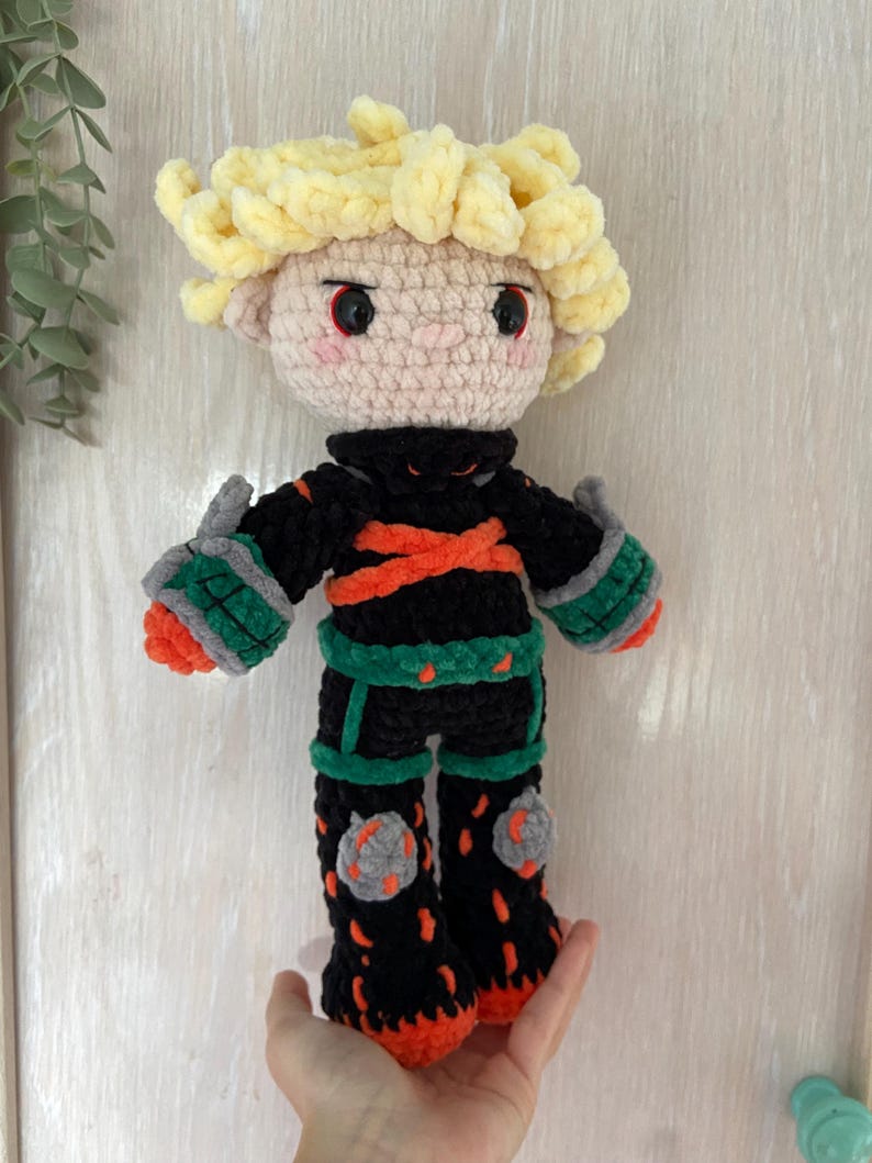 K&ouml;nnte beinhalten: Handgefertigte H&auml;kelpuppe mit einem Charakter mit stacheligen blonden Haaren, einem schwarzen Outfit mit orangefarbenen und gr&uuml;nen Akzenten und grauen Knieschonern. Die Puppe hat einen hellen Hautton und rote Augen. Die Puppe ist etwa 25 cm gro&szlig;.