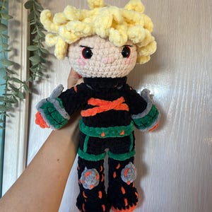 K&ouml;nnte beinhalten: Handgefertigte H&auml;kelpuppe mit einem Charakter mit stacheligen gelben Haaren, roten Augen und einem schwarzen Outfit mit orangefarbenen und gr&uuml;nen Akzenten. Die Puppe hat ein rundes Gesicht und h&auml;lt die Arme aus. Die Puppe ist etwa 30 cm gro&szlig;.