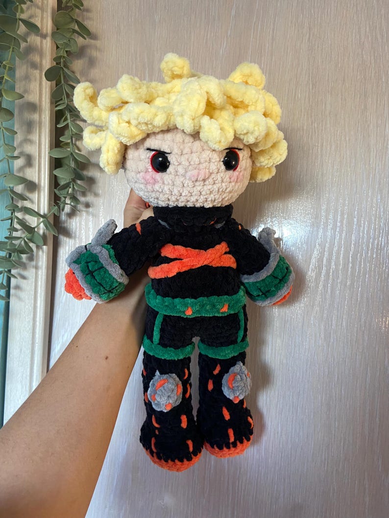 K&ouml;nnte beinhalten: Handgefertigte H&auml;kelpuppe mit einem Charakter mit stacheligen gelben Haaren, roten Augen und einem schwarzen Outfit mit orangefarbenen und gr&uuml;nen Akzenten. Die Puppe hat ein rundes Gesicht und h&auml;lt die Arme aus. Die Puppe ist etwa 30 cm gro&szlig;.