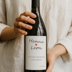 Könnte beinhalten: Eine dunkle Weinflasche mit schwarzem Verschluss und weißem Etikett. Das Etikett zeigt die Namen "Hanna" und "Leon" mit einem roten Herz, den Text "GLÜCKWUNSCH ZUR VERLOBUNG" und das Datum "12.09.2095".