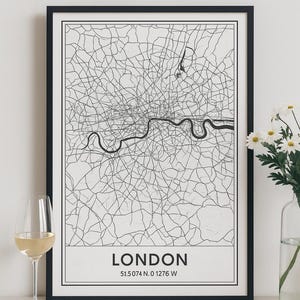 Könnte beinhalten: Gerahmte schwarz-weiße Karte von London mit der Themse. Die Karte trägt die Aufschrift "LONDON" mit Koordinaten. Ein Glas Weißwein und eine Vase mit weißen Blumen befinden sich im Vordergrund.