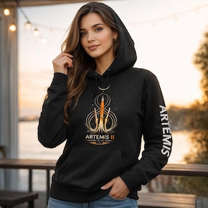 May include: Black hooded sweatshirt featuring a graphic of a rocket with the text "ARTEMIS II - MISSION TO THE MOON". The word "ARTEMIS" is printed vertically on the sleeve. The design is in shades of orange and gold.