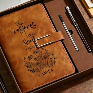 May include: A tan leather-effect journal with a metal buckle closure, featuring the text "HE restores MY soul PSALM" and a floral illustration with the name "CAMILA" printed on the cover, presented in a gift box with two black ink pens.