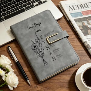 Könnte beinhalten: Ein graues Notizbuch mit einem goldfarbenen Metallverschluss. Der Einband zeigt eine schwarze Blumenillustration und den Text: God Says I am Ethan, Established, Holy, Adored, Noble. Das Notizbuch liegt auf einer Holzoberfläche neben einem Stift.