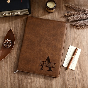 May include: A brown leather journal with the name "AMBER" and the letter "A" engraved on the front. A pen and a small piece of paper are next to the journal. The journal is closed with a zipper.