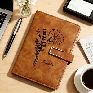 Può includere: Un diario in pelle marrone con un motivo floreale e il nome "Lydia" è esposto su una superficie di legno. Il diario ha una chiusura color oro ed è circondato da una penna, un laptop, una tazza di caffè e un piccolo vaso di fiori.