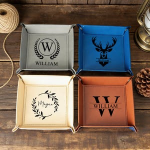 Op de afbeelding: Vier vierkante leren valet trays in grijs, blauw, crème en bruin. Elke tray heeft een gepersonaliseerd ontwerp: een letter en naam, een hertenkop, een naam in een krans en een letter en naam. De trays hebben gestikte randen.