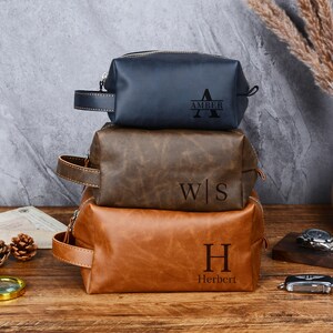 May include: Three leather wash bags in navy, brown, and tan stacked on a wooden surface. Each bag has a zip closure and a handle. The top bag is engraved with "AMBER", the middle bag with "W|S", and the bottom bag with "Herbert".