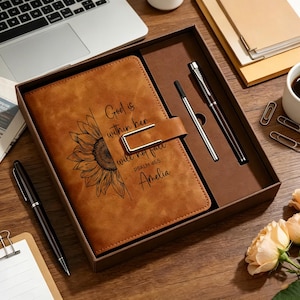 May include: A tan leather-like journal with a sunflower illustration and the text God is within her she will not fail Psalm 46:5 Amelia. The journal features a metal magnetic closure and is presented in a box with a matching black and silver pen.