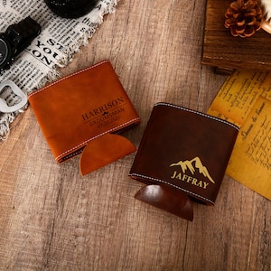 Op de afbeelding: Twee gepersonaliseerde leren drankhouders. Eén is bruin met "HARRISON GROOMSMAN 07.22.2026" en de andere is donkerbruin met "JAFFRAY" en een bergafbeelding. Beide hebben een gestikte rand.