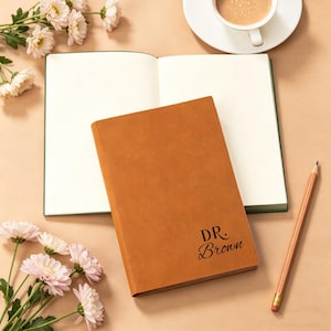 Könnte beinhalten: Ein braunes Lederjournal mit dem Namen "DR. Brown" in schwarzer Schrift. Das Journal ist geöffnet, um leere Seiten zu zeigen. Ein Bleistift und eine Tasse Kaffee befinden sich ebenfalls auf der beigen Oberfläche, zusammen mit Blumen.