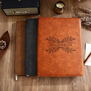 May include: Three leather journals in shades of brown and black are displayed on a wooden surface. The front journal is a light brown color and has the name "ANDERSON" engraved with a decorative leaf design. The journals are closed with zippers.