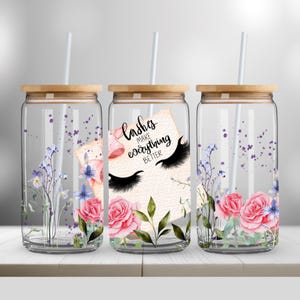 Könnte beinhalten: Drei transparente Glasbecher mit Holzdeckeln und weißen Strohhalmen. Jeder Becher hat ein Blumendesign mit rosa Rosen und lila Akzenten. Ein Becher hat die Aufschrift "lashes make everything better".