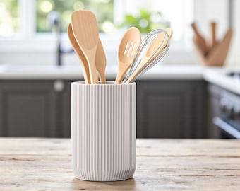 May include: A white, ribbed ceramic utensil holder on a wooden surface, filled with wooden spoons and a whisk. The kitchenware is displayed in a well-lit kitchen setting, suggesting a focus on functionality and design.