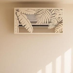 Op de afbeelding: Een crèmekleurige bank met twee bijpassende kussens, een ronde houten salontafel en een decoratieve airconditioner cover met een tropisch bladeren ontwerp. Een plant in een pot staat rechts.