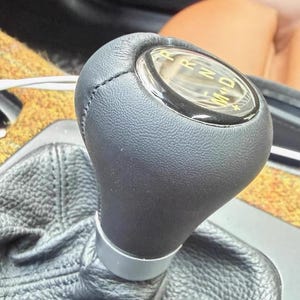 May include: A black leather gear shift knob with a textured surface and a silver base ring. The top features a glossy circular display with the letters P, R, N, D, M, and L printed in gold, indicating the transmission gear positions for the vehicle.