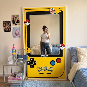 Könnte beinhalten: Ein großer rechteckiger Wandspiegel, der wie eine gelbe Game Boy Handheld-Konsole gestaltet ist. Der Rahmen zeigt Grafiken von Pikachu, Ash Ketchum und Poké Bällen. Der Text "GAME BOY" und "Pokémon" ist auf der gelben Oberfläche gedruckt.