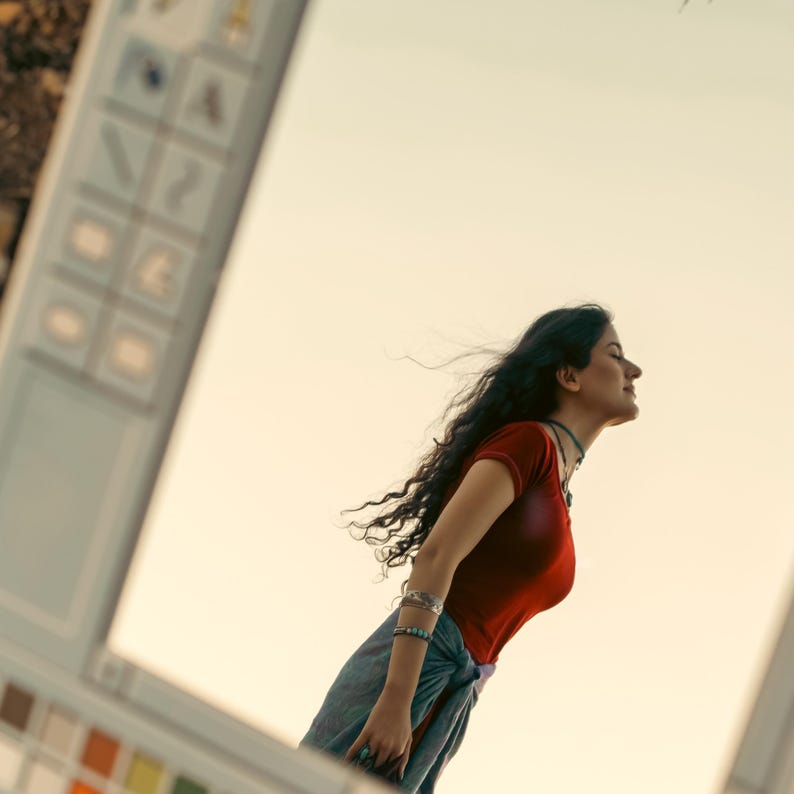 May include: A person wearing a red short-sleeved top and blue skirt, with long dark hair blowing in the wind. The person is wearing bracelets and a necklace. The background is a light yellow color.